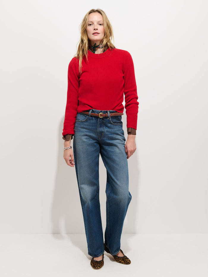 A person stands against a plain white background, wearing the Marais Sweater in Washed Cashmere—a luxurious red piece crafted from 100% cashmere—layered over a brown shirt, with blue jeans and leopard-print flats. Their shoulder-length blonde hair frames a relaxed expression, and their hands rest loosely by their sides. The extra softness of the Marais Sweater lends an elegant touch to their comfortable look.