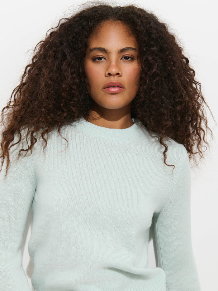 A person with long, curly hair wearing the Marais Sweater in Washed Cashmere—a light blue 100% cashmere top—stands against a plain white background, looking directly at the camera with a neutral expression.