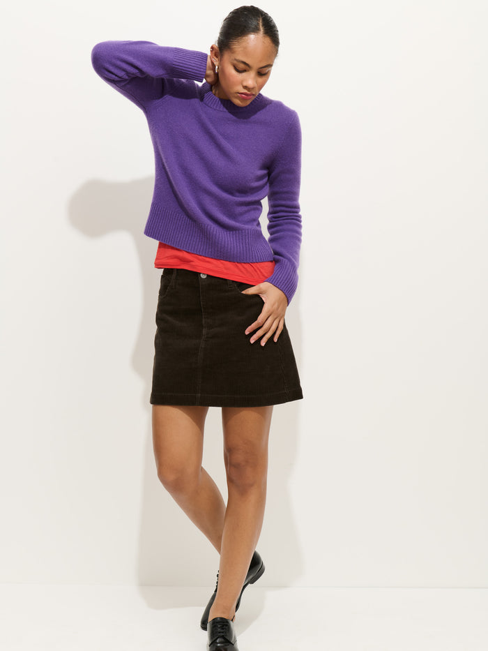 A woman stands against a white background wearing the Marais Crewneck Sweater in 100% cashmere over a red shirt, paired with a dark brown skirt and black shoes. She touches her neck and hip, gazing slightly downward.