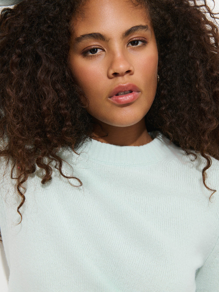 Wearing the Marais Sweater in Washed Cashmere—a light mint green luxury piece crafted from 100% cashmere—a woman with curly hair looks directly at the camera with a neutral expression.