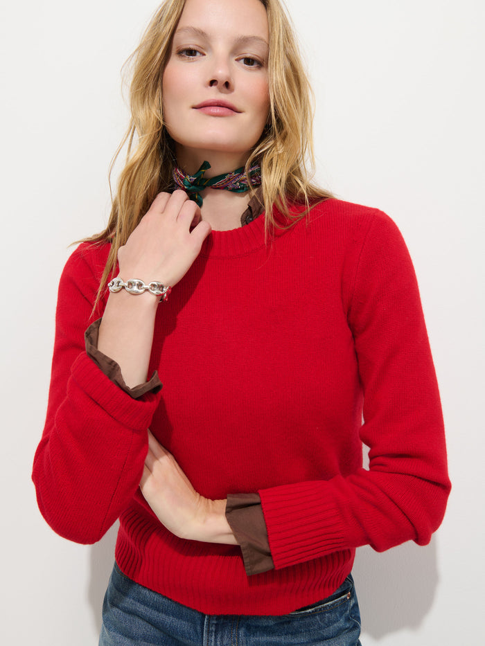 A woman with long blonde hair and a confident expression poses against a white background, exuding elegance and extra softness in the Marais Sweater in Washed Cashmere—a luxurious red piece crafted from 100% cashmere. She pairs it with blue jeans, adds a patterned scarf around her neck, and completes the look with a silver bracelet on her wrist.