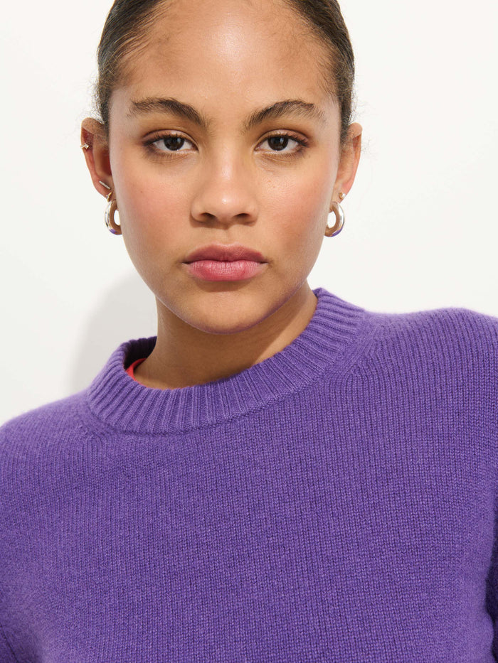 A person with pulled-back hair and gold hoop earrings wears the Marais Crewneck Sweater in 100% cashmere, purple, looking directly at the camera against a plain white background.