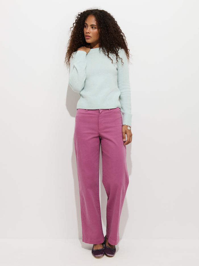 A woman with curly hair wears the Marais Sweater in Washed Cashmere—a luxurious light blue piece made from 100% cashmere for exceptional softness—paired with pink wide-leg pants and purple shoes, standing against a plain white background and gazing to her right with one hand near her neck.