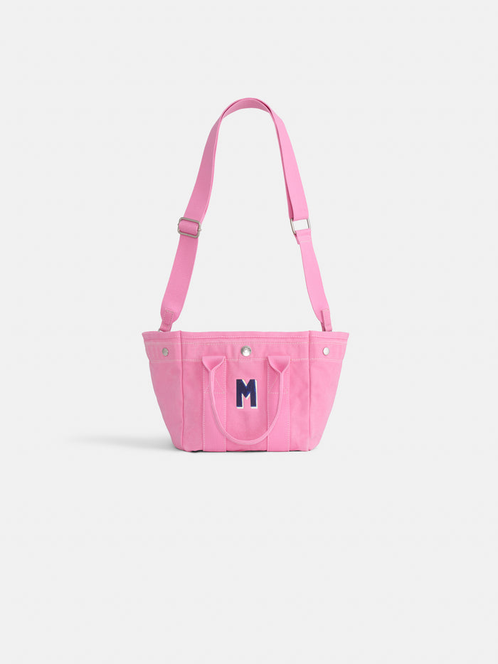 The Mini is a small pink cotton canvas tote with a front pocket, two handles, adjustable shoulder strap, and a dark blue "M" on the front—perfect for supporting breast cancer research. Shown on a plain white background.