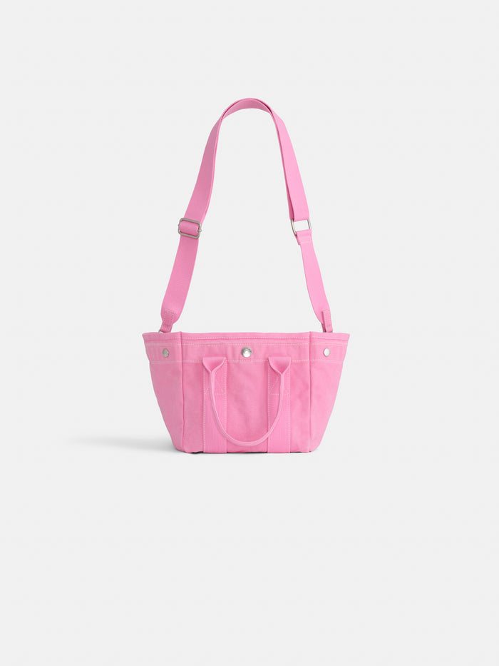 The Mini is a small, bright pink cotton canvas tote with two short handles and an adjustable shoulder strap, shown on a plain white background. Carry it in style while supporting breast cancer research.