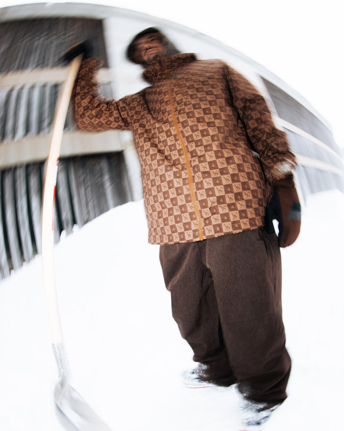 A person wearing the Volcom Men’s 2836 Insulated Jacket with a patterned brown design stands outside on snow, holding a shovel. The image is distorted with a fisheye effect, creating a warped perspective. This stylish outerwear boasts V-Science 2-Layer construction and 10K waterproof protection, making it perfect for braving wintry conditions.