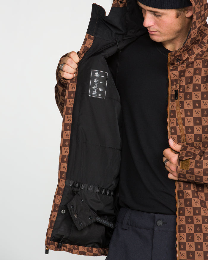A person wearing the Volcom Men’s 2836 Insulated Jacket, featuring a brown pattern and VS logos, holds it open to show the black interior and care label. Designed with V-Science 2-Layer technology for a 10K waterproof rating, they complete the look with a black shirt and black beanie.