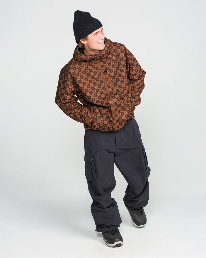 Smiling person wearing a black beanie, the Volcom Men’s 2836 Insulated Jacket in a patterned brown color, loose black cargo pants, and black sneakers stands against a plain white background.