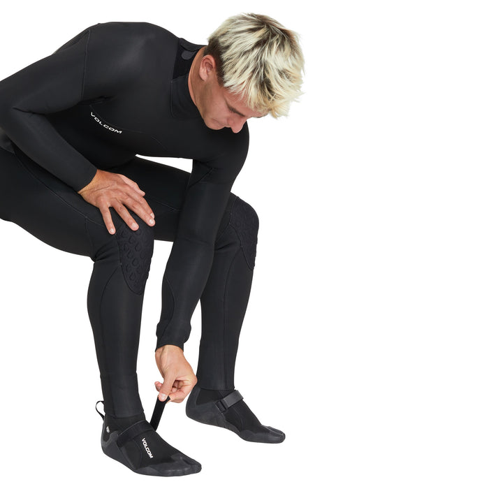 A person in a black wetsuit bends down to adjust the strap on Volcom Men's 3MM Split Toe Booties, getting ready for water activities.