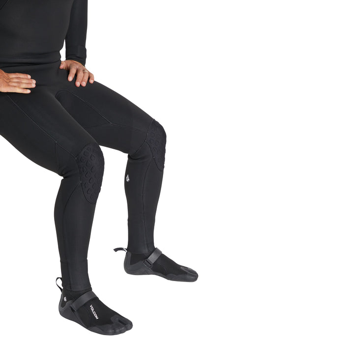 A person sits with hands on one knee, wearing Volcom Men's 3MM Split Toe Booties and a black wetsuit. The photo captures them from the chest down against a white background.