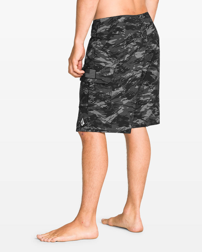 A person stands barefoot, shown from the side and back, wearing Volcom Men's 4th Of July Preemo Mod boardshorts with Cinch Fly Technology, a side pocket, and a small white logo near the hem. The background is plain white.