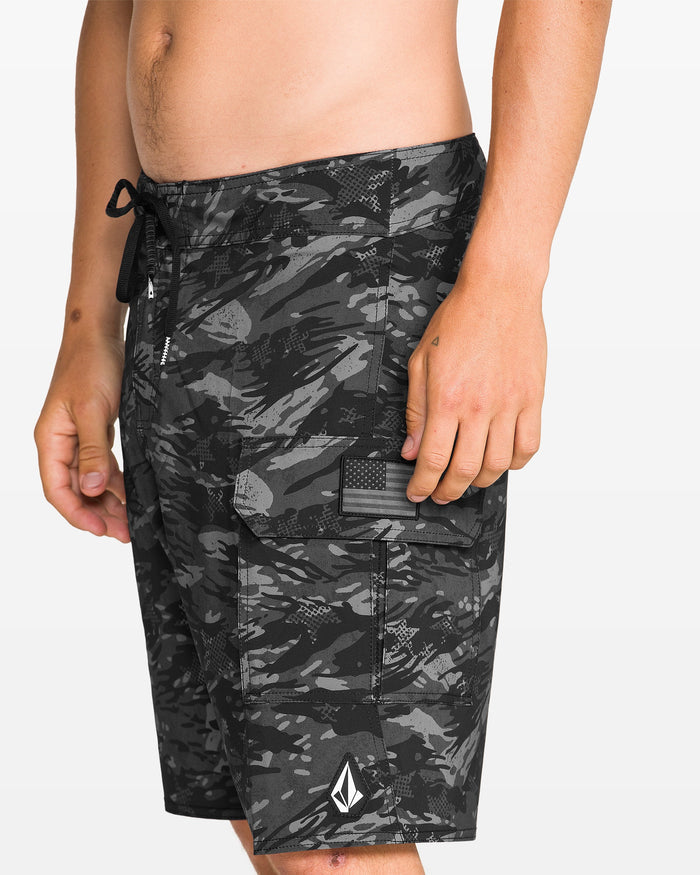 A person wearing Volcom Men's 4th Of July Preemo Mod black and gray camo boardshorts with Cinch Fly Technology, a side cargo pocket with Velcro flap, a monochrome American flag patch, and a white logo near the hem.