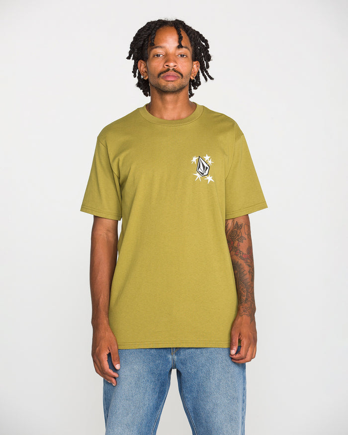 A person wearing the Volcom Men's Alive We Ride Short Sleeve Tee in light olive-green heavyweight premium cotton, featuring a soft-hand screen print graphic on the chest, and blue jeans stands against a plain white background.