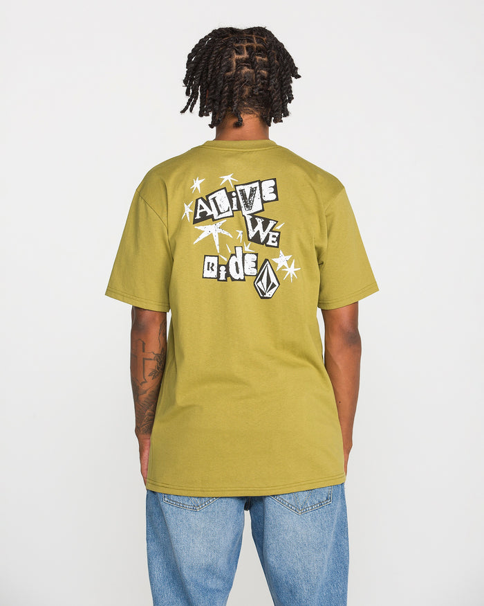 A person with short dreadlocks is shown from the back wearing the Volcom Men's Alive We Ride Short Sleeve Tee in olive green paired with blue jeans. This heavyweight premium cotton shirt features a soft-hand screen-printed graphic of shapes, a logo, and text that reads "ALIVE WE RIDE.