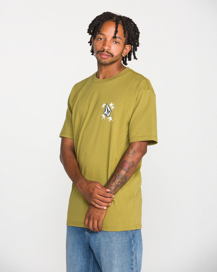 A man with short dreadlocks stands against a plain white background, looking at the camera with his hands clasped, wearing the Volcom Men's Alive We Ride Short Sleeve Tee in olive green—featuring a small white soft-hand screen print—and blue jeans.