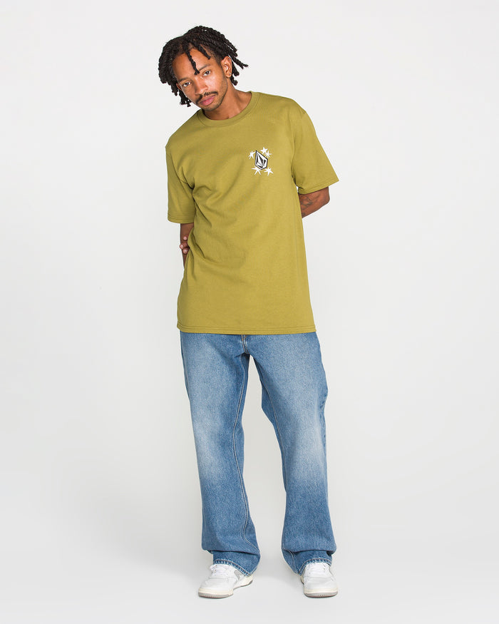 A person wearing the Volcom Men's Alive We Ride Short Sleeve Tee in yellow-green, featuring a soft-hand screen print, stands against a plain white background looking at the camera with their hands behind their back. The tee is crafted from heavyweight premium cotton for lasting comfort and style, and is paired with blue jeans and white sneakers.