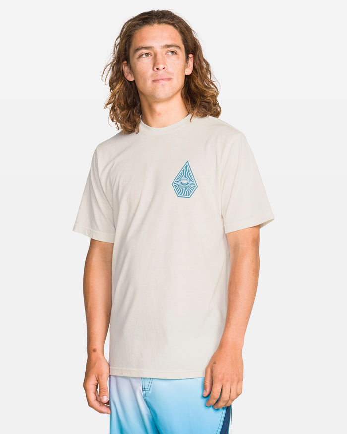A person with long brown hair is wearing the Volcom Men's All Seeing Short Sleeve Tee in Off White Heather, which has a blue geometric droplet design, and blue gradient shorts while standing against a plain white background.