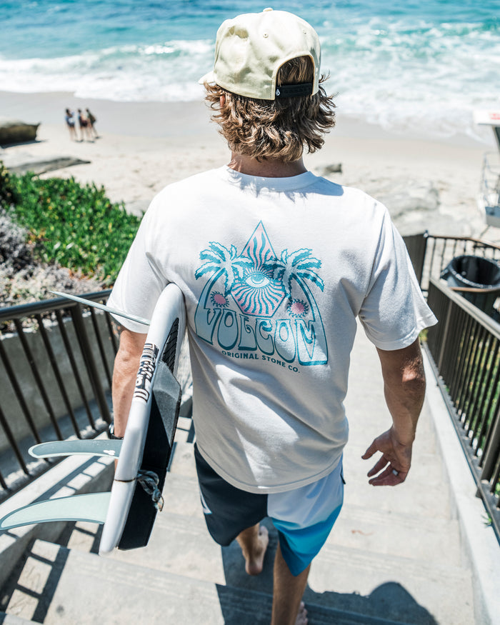 A man in a backwards cap and Volcom Men's All Seeing Short Sleeve Tee carries a surfboard down stairs toward a sandy beach and blue ocean on a sunny day.