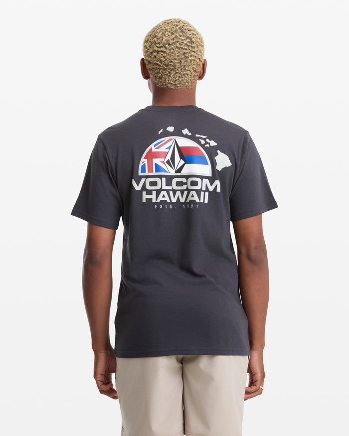 A man with short blond hair wears the Volcom Men's Allegiance Short Sleeve Tee, featuring "VOLCOM HAWAII" text, a Hawaii outline, and flag graphic on the back, standing against a plain white background.
