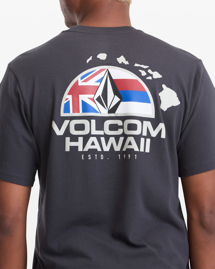 A person wearing the Volcom Men's Allegiance Short Sleeve Tee, a black T-shirt featuring "VOLCOM HAWAII ESTD. 1991," a graphic of the Hawaiian islands, stylized logo, and a partial Hawaiian flag on the back.