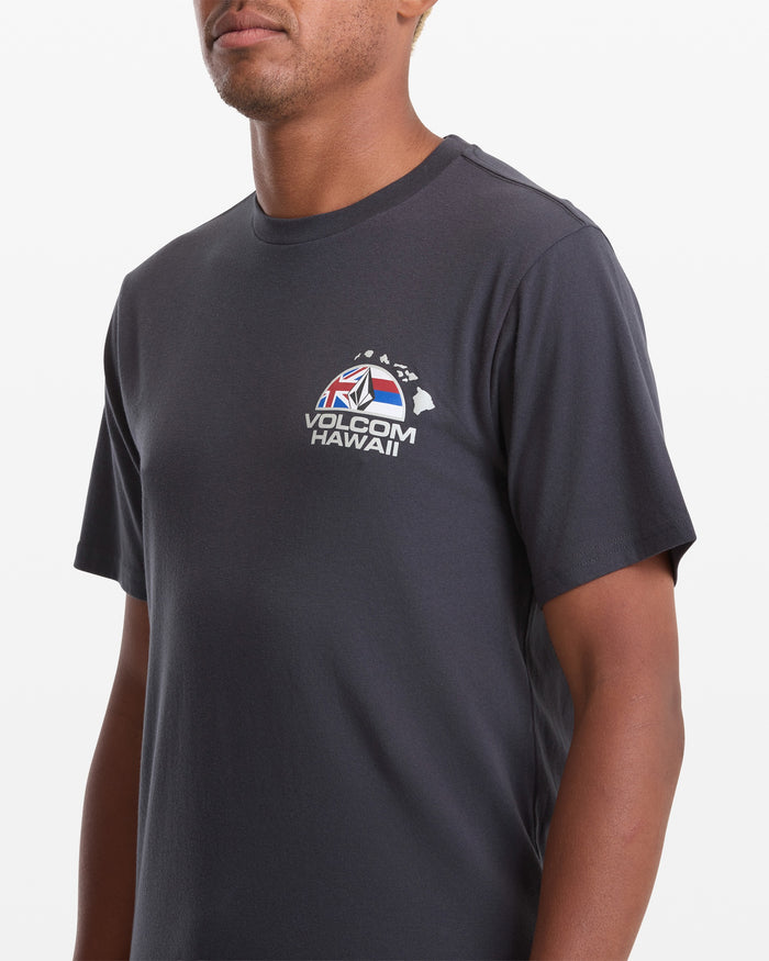 A man wears a dark grey Volcom Men's Allegiance Short Sleeve Tee with a small red, white, and blue logo and Hawaiian islands outline on the left chest.