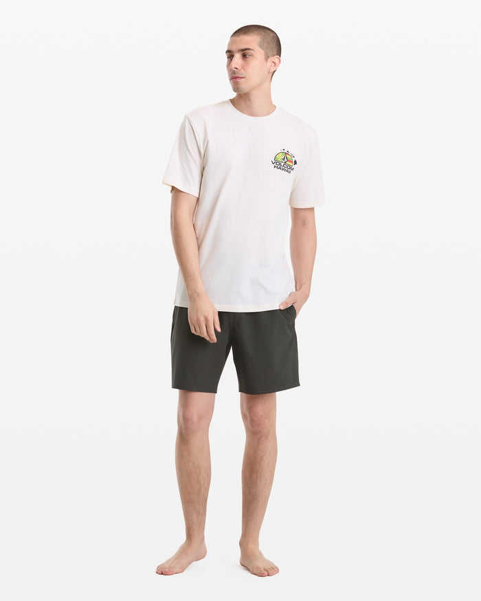 A barefoot man stands against a white background wearing the Volcom Men's Allegiance Short Sleeve Tee with a small colorful chest graphic and dark shorts, one hand in his pocket, looking to the side.