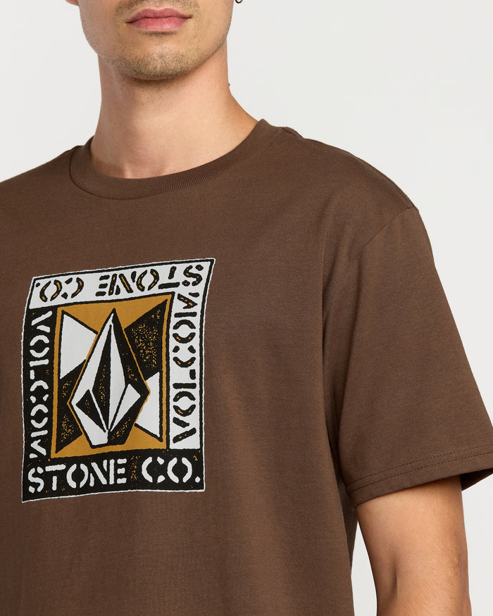 A man wears the Volcom Men's Angle Iron Short Sleeve Tee in brown, featuring a geometric logo and "VOLCOM STONE CO." on the chest. Only his lower face and upper torso show. Made from premium ring-spun cotton for comfort.