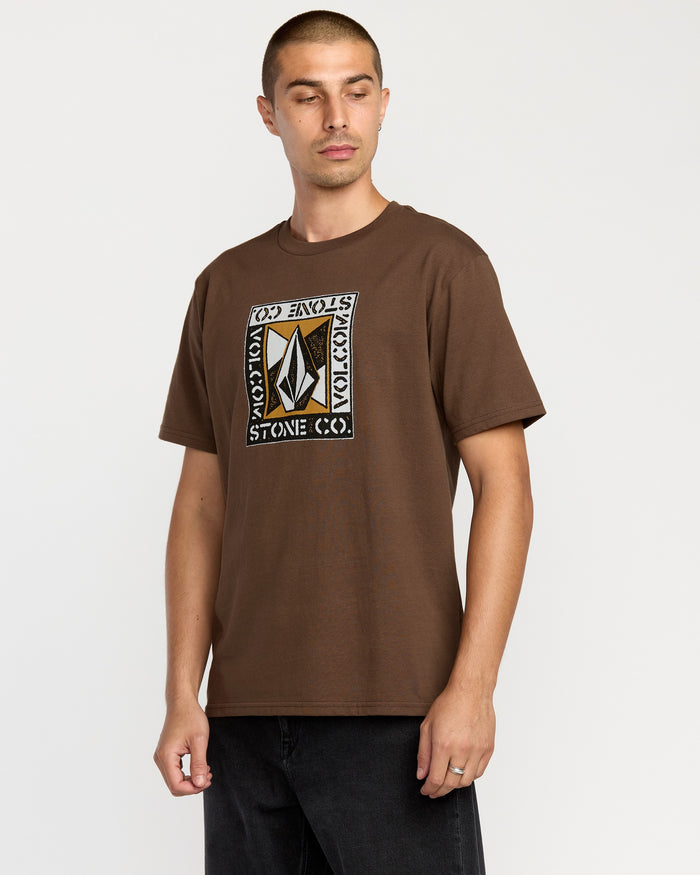 A man wears Volcom's Men's Angle Iron Short Sleeve Tee, a brown premium ring-spun cotton T-shirt featuring a geometric logo and the words VOLCOM STONE CO., standing against a plain white background.