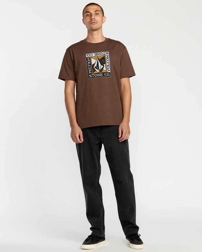 A man stands against a plain white background wearing the Volcom Men's Angle Iron Short Sleeve Tee, paired with black jeans and black sneakers. He has short hair, a neutral expression, and his arms are relaxed by his sides.