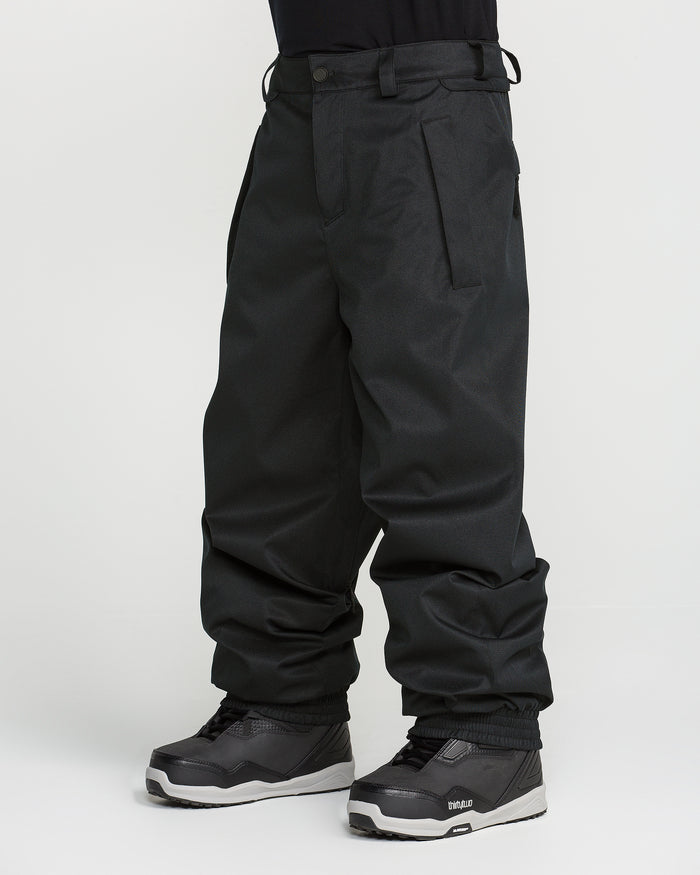 A person wearing Volcom Men’s Arthur 20K Pants in loose-fitting black recycled twill and black snowboard boots, standing against a plain white background.