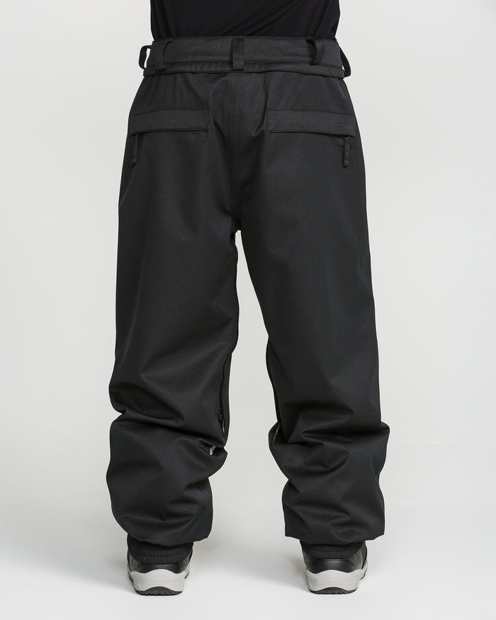 A person wearing loose-fitting black Volcom Men’s Arthur 20K Pants, featuring zipper pockets and crafted from recycled twill, paired with white sneakers and photographed from the waist down against a plain white background.