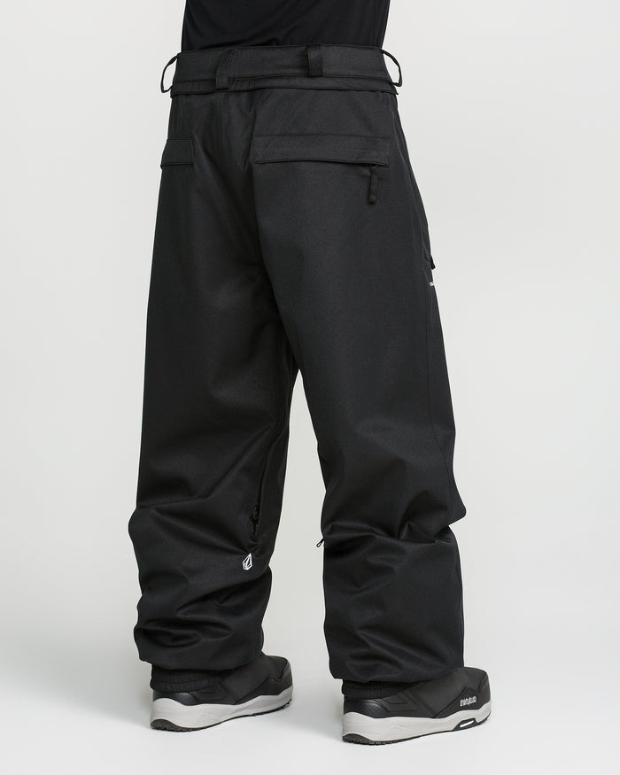 A person is seen from behind wearing loose-fitting black Volcom Men’s Arthur 20K Pants, crafted from recycled twill, paired with black sneakers against a plain white background. The pants feature zippered pockets and belt loops, and their hems drape over most of the shoes.
