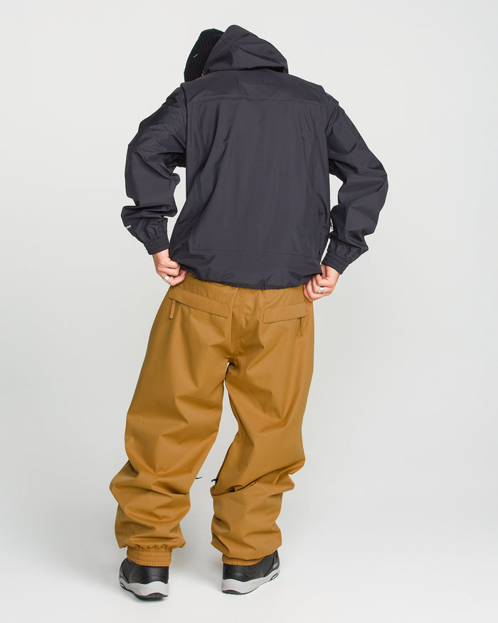 A person facing away, wearing a black hooded jacket and loose brown Volcom Men’s Arthur 20K Pants crafted from recycled twill with V-Science 2-Layer technology, paired with black and white sneakers, stands against a plain white background.