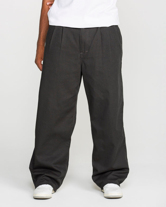 A person, visible from the waist down, stands against a plain white background wearing loose-fitting black Men's Baggos Pleat Pants by Volcom, paired with a white shirt and white sneakers.