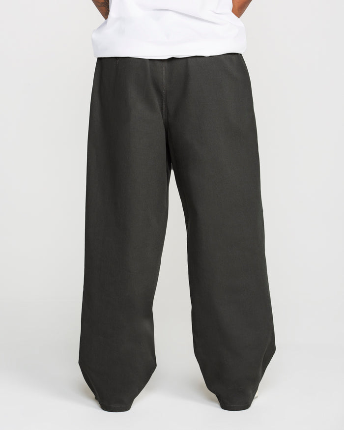 A person models the Volcom Men's Baggos Pleat Pants—a pair of loose-fitting, wide-leg black pants—paired with a white shirt, standing with their hands behind their back against a plain white background.