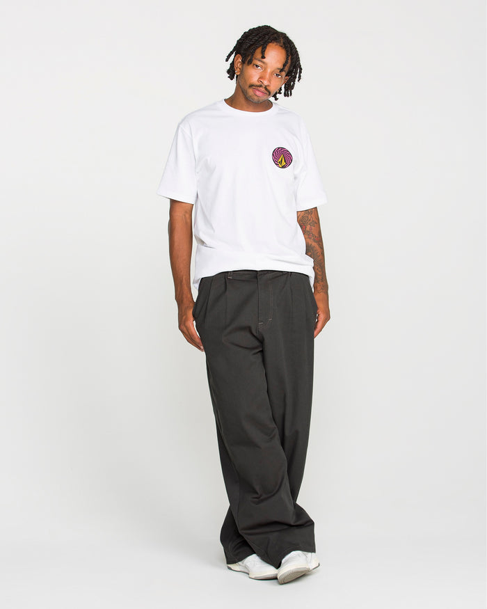A man stands against a plain white background wearing a white t-shirt with a small circular logo, Volcom Men's Baggos Pleat Pants in black, and white shoes.