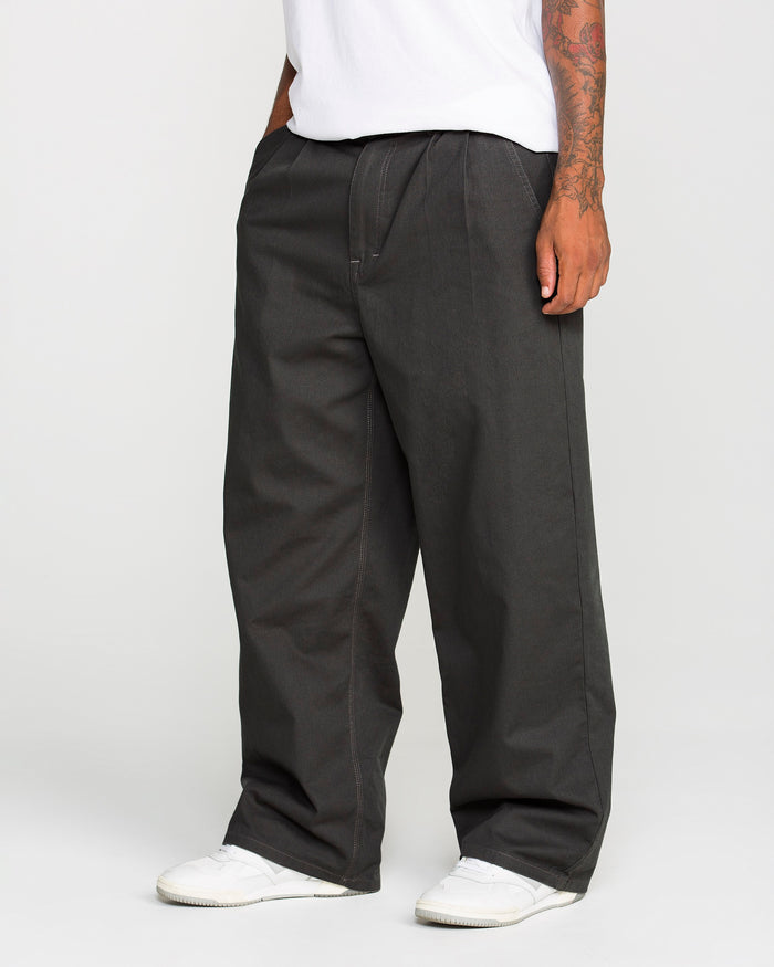 A person is seen from the waist down wearing Volcom Men's Baggos Pleat Pants in a loose, baggy fit styled like chinos. The outfit is paired with white sneakers and a white T-shirt, showing tattoos on their right forearm. The plain, light-colored background reflects a relaxed Volcom aesthetic.