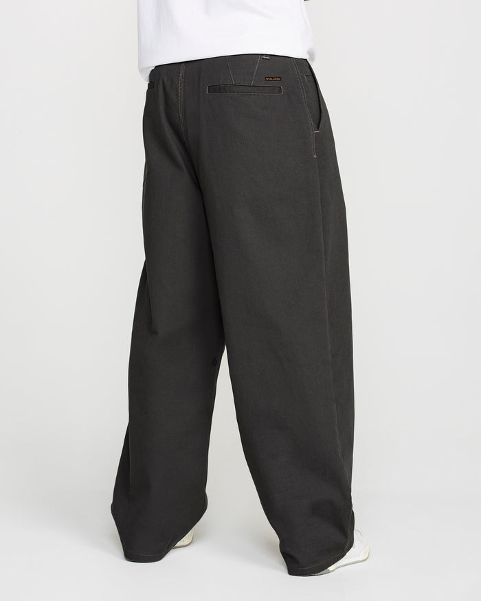 A person wearing Volcom Men's Baggos Pleat Pants in black, styled with a white shirt, is shown from the back. The loose-fitting pants feature a straight leg cut and pockets. Only the lower body is visible against a plain white background.