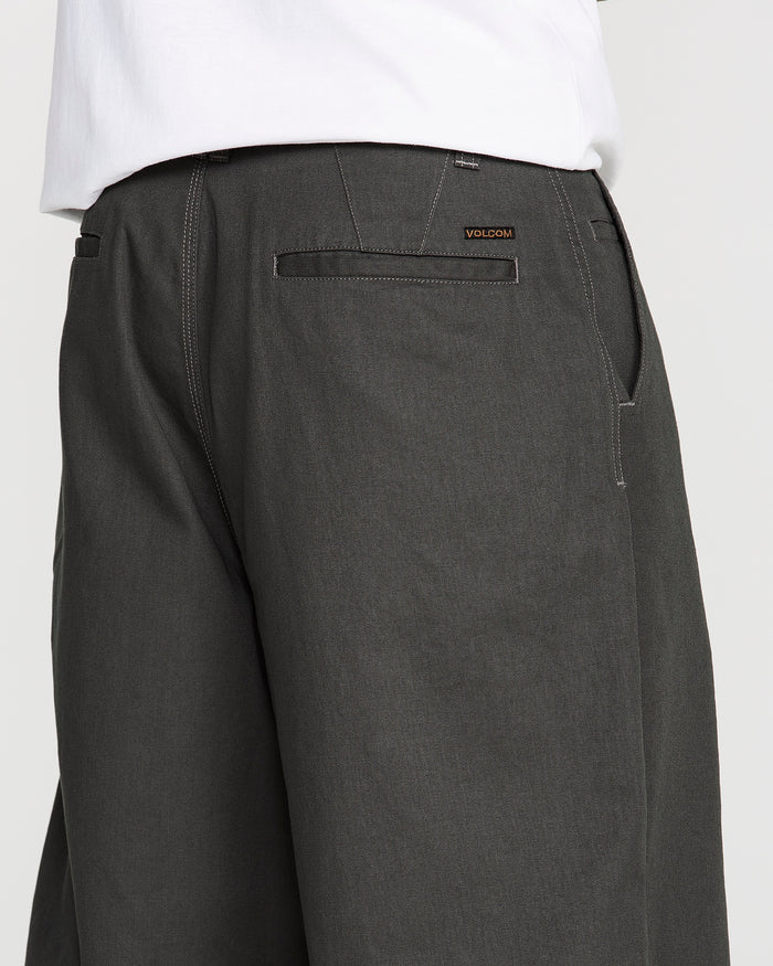 A person is wearing Volcom Men's Baggos Pleat Pants in dark gray cotton with a white shirt, shown from the waist down at an angle so the back pocket and Volcom brand label are visible.