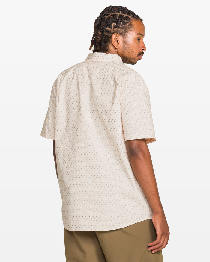 A man with short dreadlocks models the Volcom Men's Bankstone II Short Sleeve Tee, paired with olive green pants, standing turned away and looking over his shoulder against a plain white background.