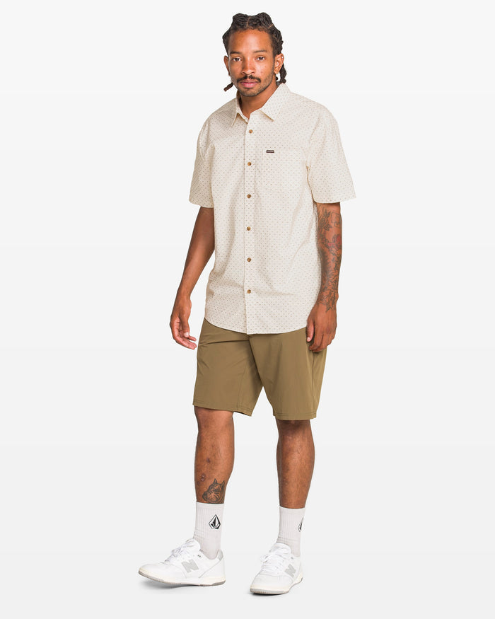 A man with braided hair wears the Volcom Men's Bankstone II Short Sleeve Tee, olive green shorts, white socks, and white sneakers as he stands against a plain white background.