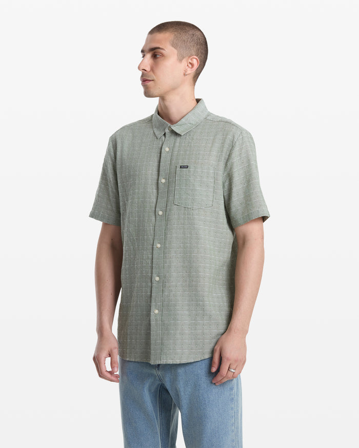 A man with short hair wears the Volcom Men's Bankstone II Short Sleeve shirt in light green with a chest pocket and yarn-dyed dobby texture, paired with light blue jeans, standing against a plain white background.