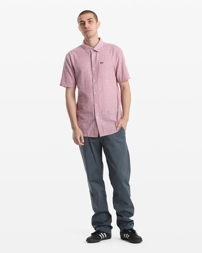 A man with short hair and a neutral expression stands against a white background wearing the Volcom Men's Bankstone II Short Sleeve shirt, blue-gray pants, and black Adidas sneakers with white stripes, one hand in his pocket.