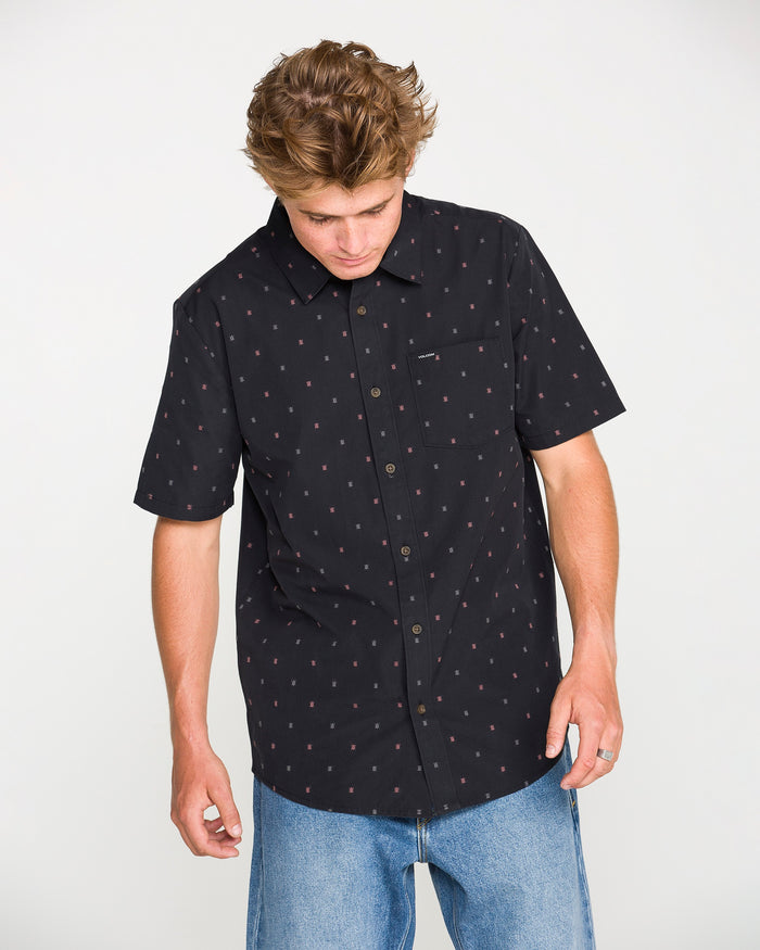 A person wearing the Volcom Men's Bankstone Short Sleeve Shirt paired with light blue jeans looks down with both hands at their sides against a plain background.