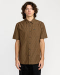 A man with wavy brown hair wears the Volcom Men's Bankstone Short Sleeve Shirt, a brown textured dobby classic-fit shirt with a small print pattern and chest pocket, standing against a plain white background.