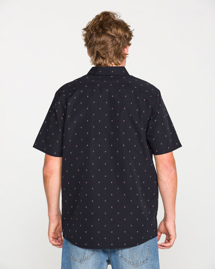 A person with light brown hair is shown from the back against a plain white background, wearing the Volcom Men's Bankstone Short Sleeve Shirt—a black button-up featuring a small red dotted pattern—paired with blue jeans.