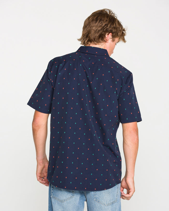A person with light brown hair, wearing the Volcom Men's Bankstone Short Sleeve Shirt in classic fit navy blue with a small colorful dot pattern in a dobby weave, paired with light blue jeans, is facing away from the camera against a plain white background.