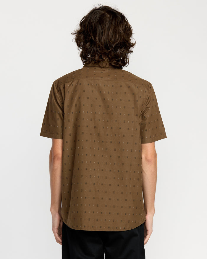 A person with medium-length wavy brown hair is shown from the back, wearing the Volcom Men's Bankstone Short Sleeve Shirt in classic fit and black pants, standing against a plain white background.