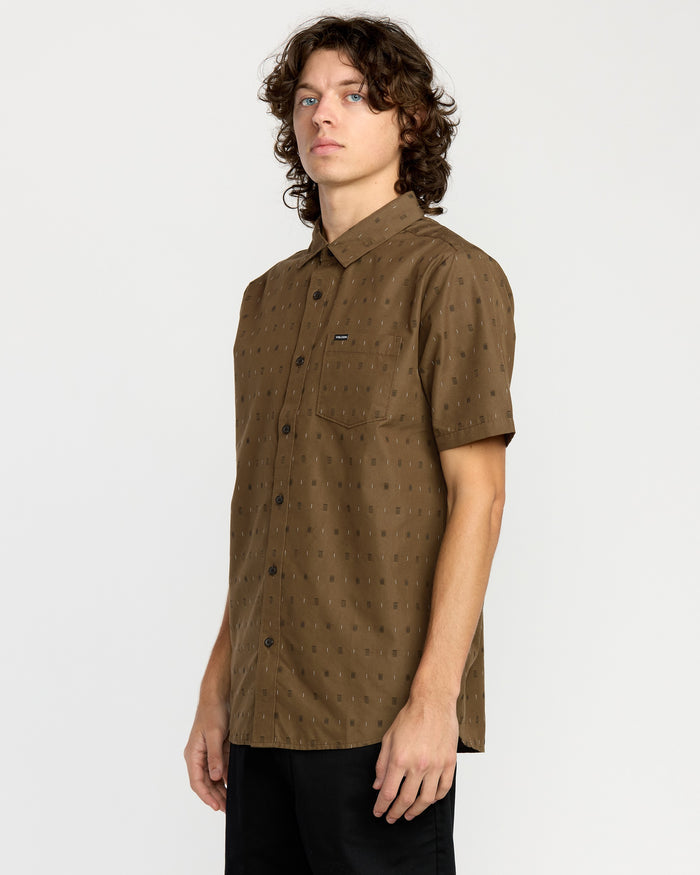 A young man with curly hair stands sideways against a plain light background, wearing the Volcom Men's Bankstone Short Sleeve Shirt in a classic fit with a subtle dotted pattern, paired with black pants.