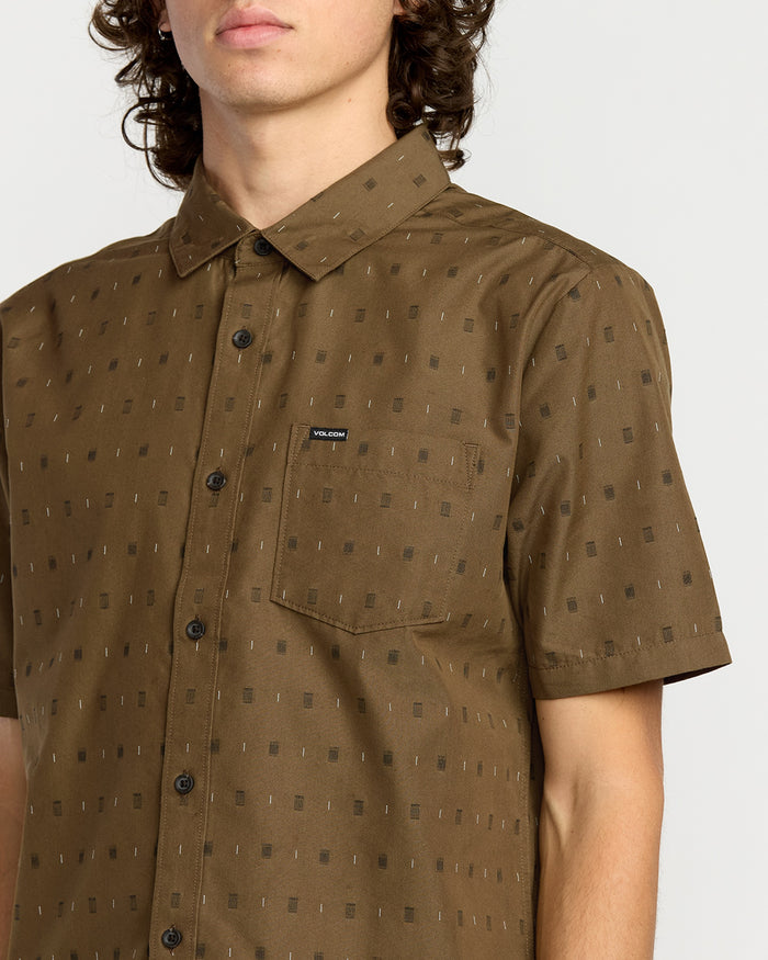 A man wears the Volcom Men's Bankstone Short Sleeve Shirt in brown, featuring a classic fit, subtle geometric pattern, chest pocket with a Volcom label, and short sleeves. His face is partially out of frame.